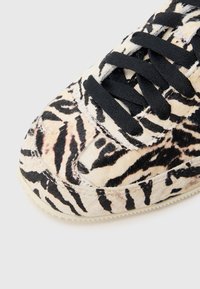 Nízký sportovní boty s potiskem tygřích pruhů v krémové a černé barvě, vyrobené z texturovaného materiálu, s černými tkaničkami a gumovou podrážkou.