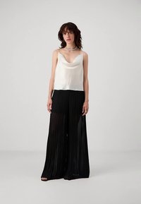 Kvinde iført en hvid satin top og sorte gennemsigtige plisserede bredbukser, der står mod en ensfarvet grå baggrund.