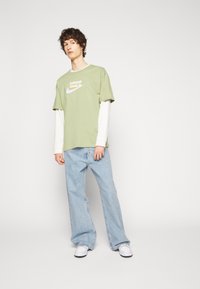 T-shirt en coton vert avec le logo "Nike", superposé à une chemise à manches longues blanche, associé à un jean large bleu clair et à des baskets blanches.