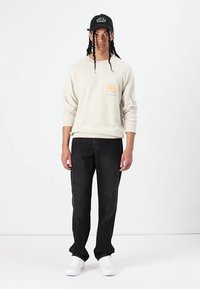 Cremfarbener Sweatshirt mit einem kleinen orangen Sonnenmotiv auf der Brust, kombiniert mit schwarzen Jeans und weißen Sneakers. Schwarze Kappe mit weißem Text.