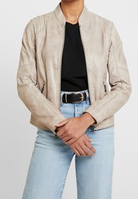 Béžová umělá kožená bunda s vysokým límcem, předním zipem, bočními kapsami a jemným žebrováním na ramenech. Nositá přes černý top.