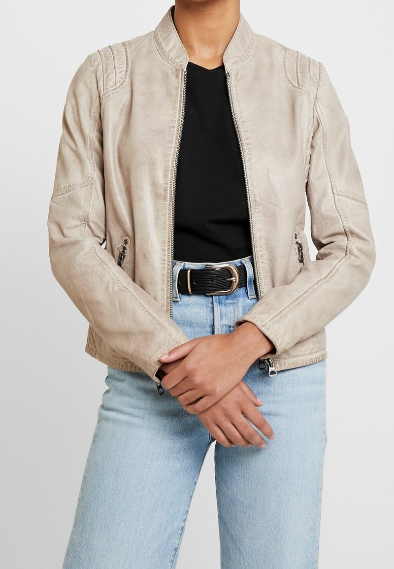 Béžová umělá kožená bunda s vysokým límcem, předním zipem, bočními kapsami a jemným žebrováním na ramenech. Nositá přes černý top.