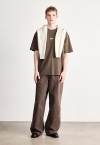 Brązowy, luźny t-shirt z małym logo, zestawiony z szerokimi brązowymi spodniami. Kremowy prążkowany sweter zarzucony na ramiona. Czarne buty.