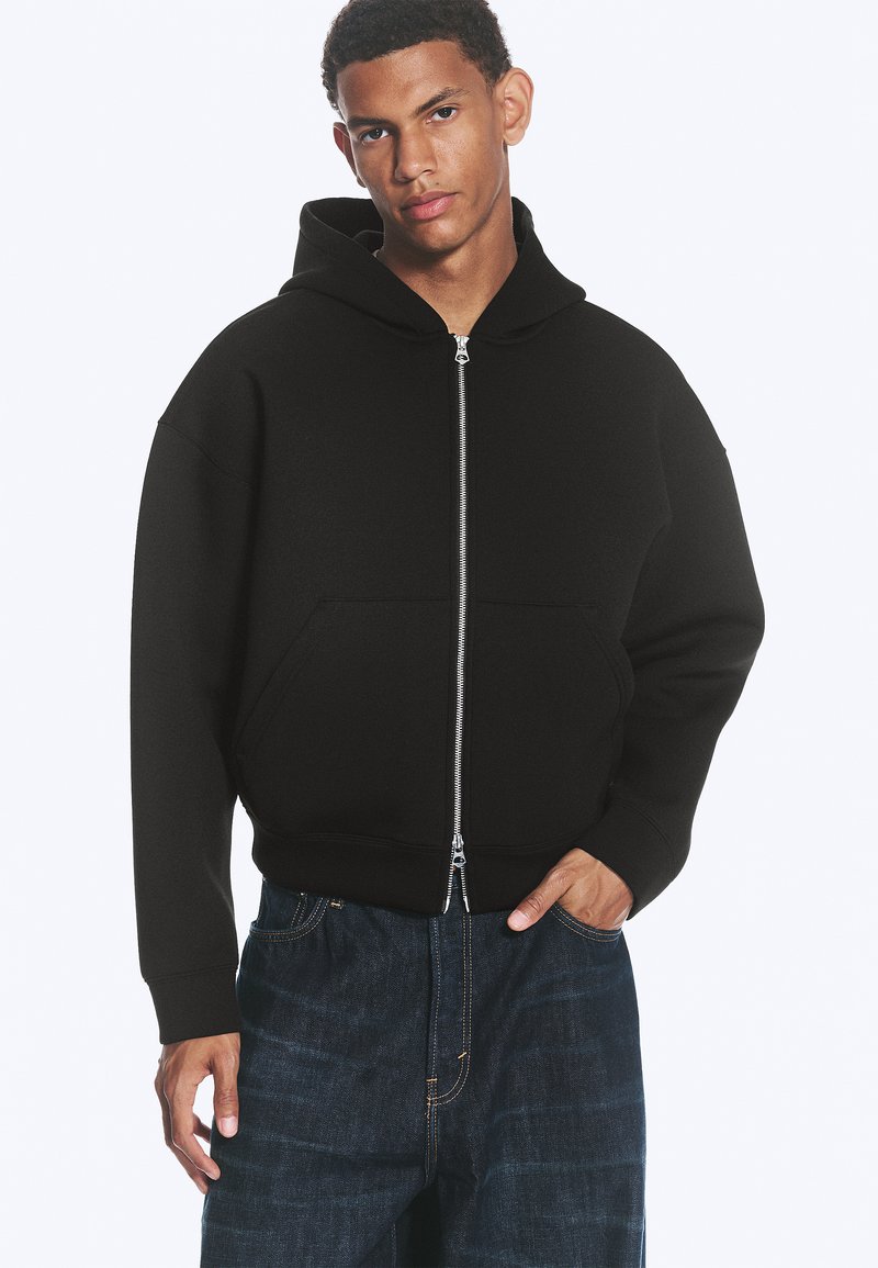 Jeune homme portant un sweat à capuche noir zippé avec une poche avant et un jean bleu foncé, debout avec une main dans la poche.