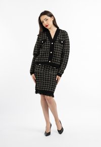 Completo in tweed nero e crema, con giacca cropped dotata di grandi bottoni e tasche, abbinata a una gonna a frange di lunghezza al ginocchio.