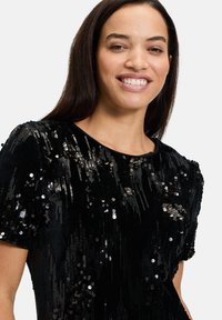 Czarny top z cekinami, krótki rękaw, o miękkiej teksturze i okrągłym dekolcie. Cekiny tworzą połyskujący efekt na materiale.