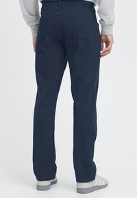 Temno modre hlače iz bombažnega blaga, z ravnim krojem, s dvema zadnjima žepoma, nošene s sivimi čevlji. Gladek material, minimalističen dizajn.