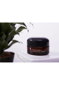 Bocal en verre brun avec un couvercle noir étiqueté "Crème Contour des Yeux Réparatrice Multi-Fonction au Snail", surface lisse, forme ronde.