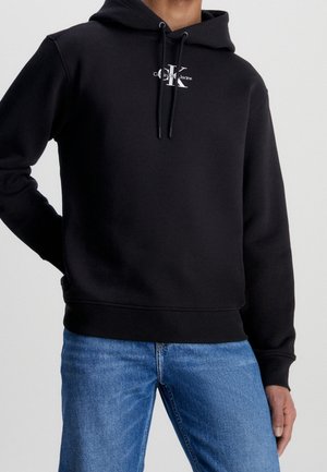 Osoba ubrana w czarną bluzę z kapturem Calvin Klein z logo na piersi oraz niebieskie dżinsy, stojąca na jednolitym jasnym tle.