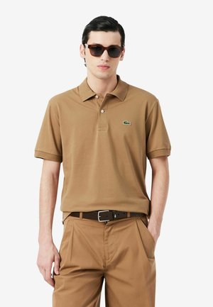 Uomo che indossa una polo marrone con il logo del coccodrillo verde, pantaloni marroni abbinati, cintura nera e occhiali da sole scuri, in piedi con una mano in tasca.