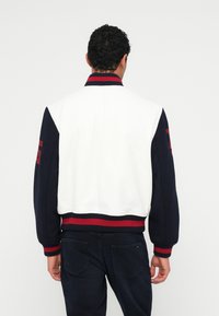 Giacca bomber bianca e blu navy con collo e polsini a coste, con dettagli rossi. Design semplice con tessuto testurizzato e un aspetto sportivo.
