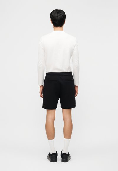 Άνδρας στέκεται γυρισμένος με την πλάτη, φοράει λευκό πουκάμισο με μακριά μανίκια, μαύρα σορτς, λευκές κάλτσες και μαύρα παπούτσια σε απλό φόντο.