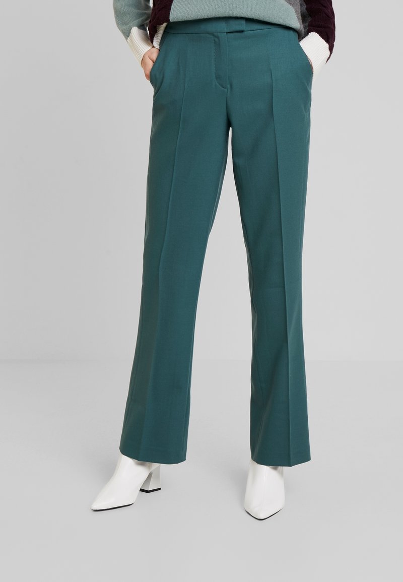 Vrouw in een teal op maat gemaakte broek met voortassen, gecombineerd met een veelkleurige trui en witte enkellaarsjes met hakken, staand op een grijze vloer.