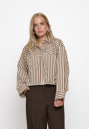Junge Frau mit langen blonden Haaren, die ein braun-weiß vertikal gestreiftes Hemd und dunkelbraune Hose trägt, steht vor einem schlichten weißen Hintergrund.