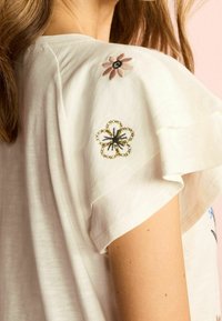 Camisa blanca, ligera, de manga corta. Presenta bordado floral en rosa, negro y azul en los hombros, textura suave y corte relajado.