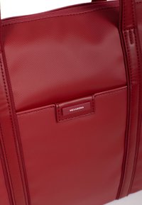 Czerwona torba tote z teksturowaną powierzchnią, przednią kieszenią z metką oraz solidnymi uchwytami. Prostokątny kształt, minimalistyczny design.