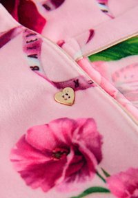 Rosa Fleece-Stoff mit Blumenmuster, mit einem kleinen goldenen herzförmigen Logodetail nahe dem Reißverschluss. Weicher Textur, lebendige Farben.