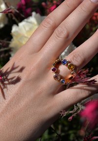 Gold ring with seven colorful gemstones (pink, orange, blue, and purple) set in a circular pattern on a finger against a floral background.