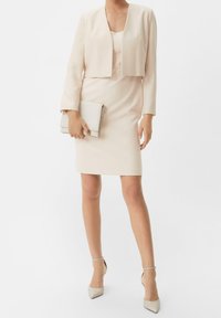 Blekt rosa klänning med figurnära siluett och knälång längd, kombinerad med en kort jacka och en liten ljusgrå kuvertväska.