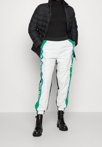 Vita träningsbyxor med gröna och blå sidostråkar, i kombination med en svart puffjacka och svarta schelgöringskängor.