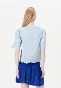 Ανοιχτού μπλε πουκάμισο με κοντά φουσκωτά μανίκια, που διαθέτει κεντητή στο κάτω μέρος και λεπτομέρειες από μάτι, σε συνδυασμό με μια υφασμάτινη μπλε φούστα.