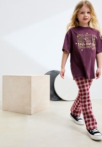 T-shirt violet oversize avec texte jaune et étoiles, associée à un pantalon à carreaux rouges et blancs. Baskets noires. Fond de couleur claire.