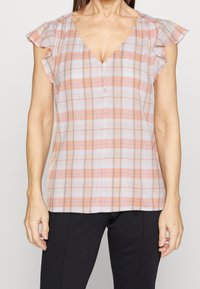 Blouse légère à carreaux avec un col en V, des manches courtes volantées et des boutons à l'avant. Les couleurs comprennent des tons de rose pâle, blanc et gris.