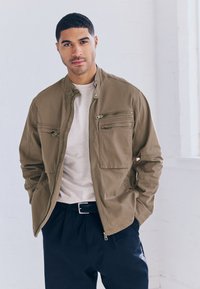 Kaki suknjič z zadrgo in visokim ovratnikom, sprednjimi žepi in gladko teksturo, kombiniran z svetlo sivim T-shirtom in črnimi hlačami.