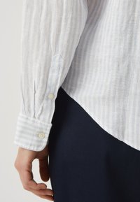 Jasnoniebieska w białe paski koszula z lnu, z guzikami przy mankietach i zaokrąglonym dołem. Proste granatowe spodnie częściowo widoczne.