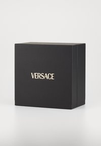 Caja de cartón negra con un acabado texturizado, con el logo dorado en relieve "VERSACE" en la parte frontal. Diseño rectangular simple.