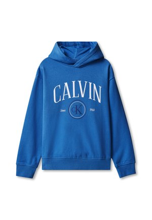 Chłopiec ubrany w niebieską bluzę z kapturem Calvin Klein, szare szerokie jeansy i białe trampki, stoi na jednolitym tle.