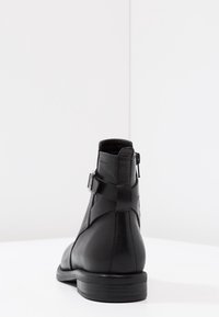 Botines de cuero negro que presentan una textura suave, un cierre lateral con cremallera y un acento de correa cerca del tobillo. Suela de goma plana.