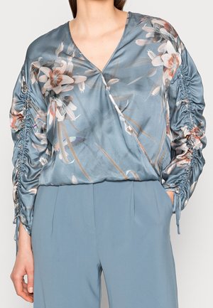 Nainen pukeutuneena silkkiseen siniseen kukkaiseen kietaisupaitaan, jossa on poimutetut pitkät hihat, ja korkeavyötäröisiin sinisiin housuihin, käsi taskussa, neutraalia taustaa vasten.