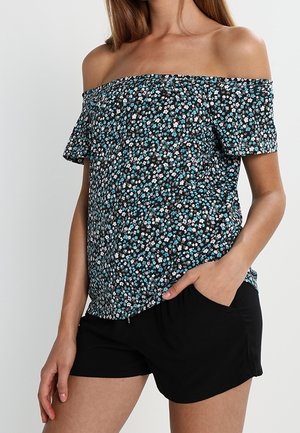 Frau trägt ein schwarzes schulterfreies Oberteil mit kleinen blauen und weißen Blumen und schwarze Shorts, eine Hand in der Tasche.