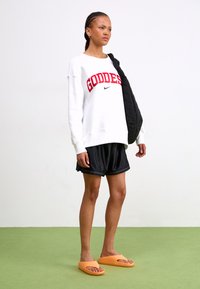 Fehér pulóver piros "GODDESS" felirattal és Nike logóval, fekete sportos rövidnadrággal és narancssárga papuccsal párosítva. Fekete steppelt táska a vállán.