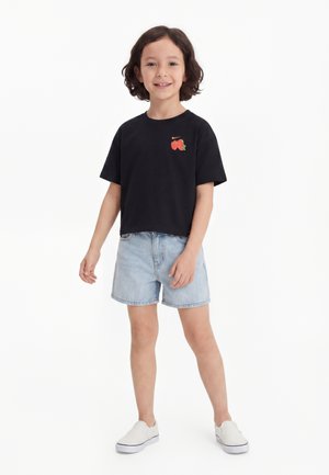 Barn med mørkt krøllet hår iført en sort T-shirt med frugtmotiv, lyse denimshorts og hvide slip-on sko, stående på hvid baggrund.
