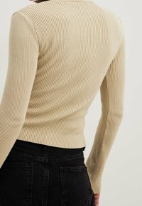 Haut à manches longues beige côtelé porté avec un jean noir taille haute, vu de dos avec le bras gauche de la personne légèrement fléchi.