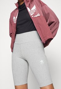 Mörkröd zip-up jacka med vit logga, svart åtsittande topp och högmidjade grå cykelbyxor med liten vit logga. Släta tygstrukturer.