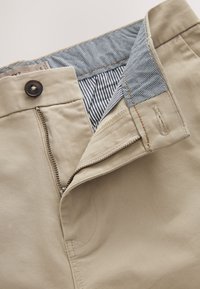 Pantalon en coton beige avec fermeture éclair partiellement ouverte, bouton noir et ceinture intérieure montrant une doublure rayée et à pois.