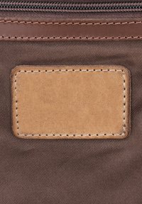 Écusson rectangulaire en cuir beige avec coutures sur fond en tissu marron sous une fermeture éclair avec garniture en cuir.