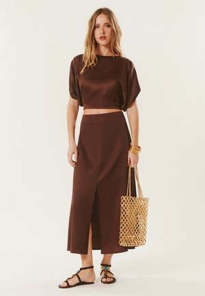 Femme portant un crop top marron et une jupe taille haute avec une fente devant, tenant un sac tressé beige, et portant des sandales plates noires et des bracelets dorés.