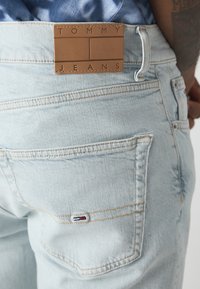 Světle modré denimové džíny s koženou nášivkou označenou "TOMMY JEANS" v pase, s jemnými švy a malou logovou nálepkou na kapse.