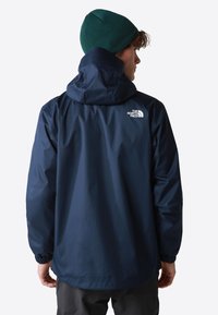 Chaqueta impermeable de color azul marino con capucha, puños elásticos y un logo blanco en la parte posterior. Presenta una textura suave y mate.