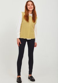 Light green knitted vest over a white collared blouse, paired with dark skinny jeans and black loafers, creating a layered look.