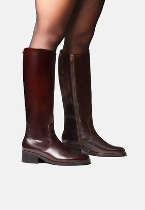 Bottes en cuir brun foncé brillantes montant jusqu'aux genoux avec des talons bas, portées avec des collants noirs transparents, montrées sous angles latéral et frontal sur fond uni.
