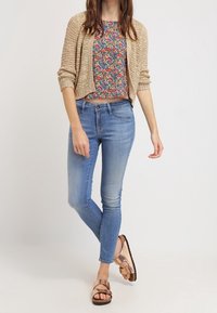 Beige stickad cardigan över en blommig kort topp, ihop med ljusblå skinny jeans och bronsfärgade sandaler. Enkel design, avslappnad passform.