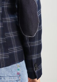 Manche de veste à carreaux bleu marine avec un patch coude en denim foncé et deux boutons noirs, portée sur un jean bleu clair avec une broderie colorée.