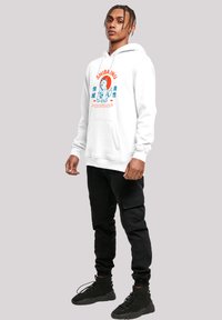 White hoodie with Shiba Inu graphic, blue and orange text, kangaroo pocket, and cuffed sleeves, paired with black cargo pants and black sneakers.
