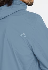 Ljusblå vattentät jacka med huva, med en diskret logotyp på axeln och slät yta, framhävd av rena sömmar.