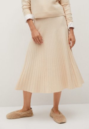 Falda plisada beige hecha de tela suave, de longitud media, con una superficie texturizada. Combinada con pantuflas beige esponjosas.